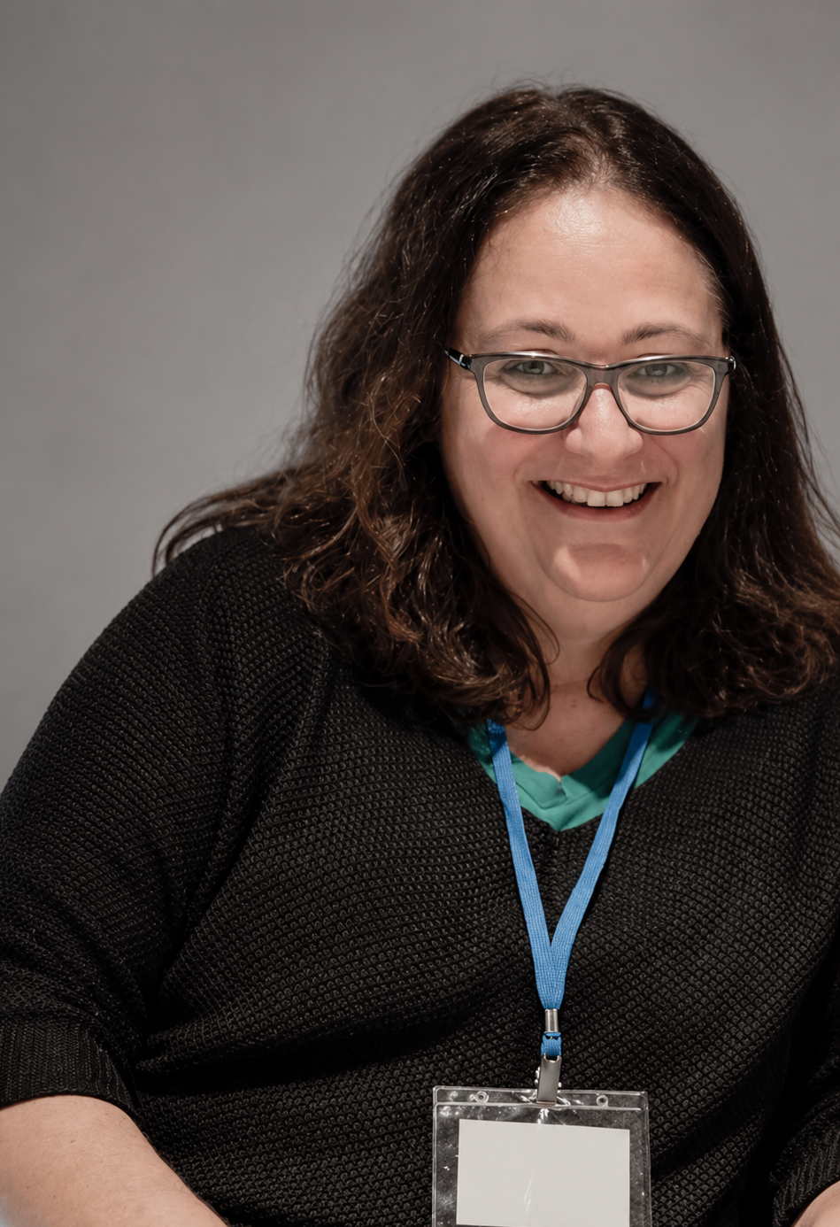 A smiling woman with glasses and dark, wavy shoulder-length hair, wearing a black sweater and an ID badge on a blue lanyard, seated at a white table against a gray background.