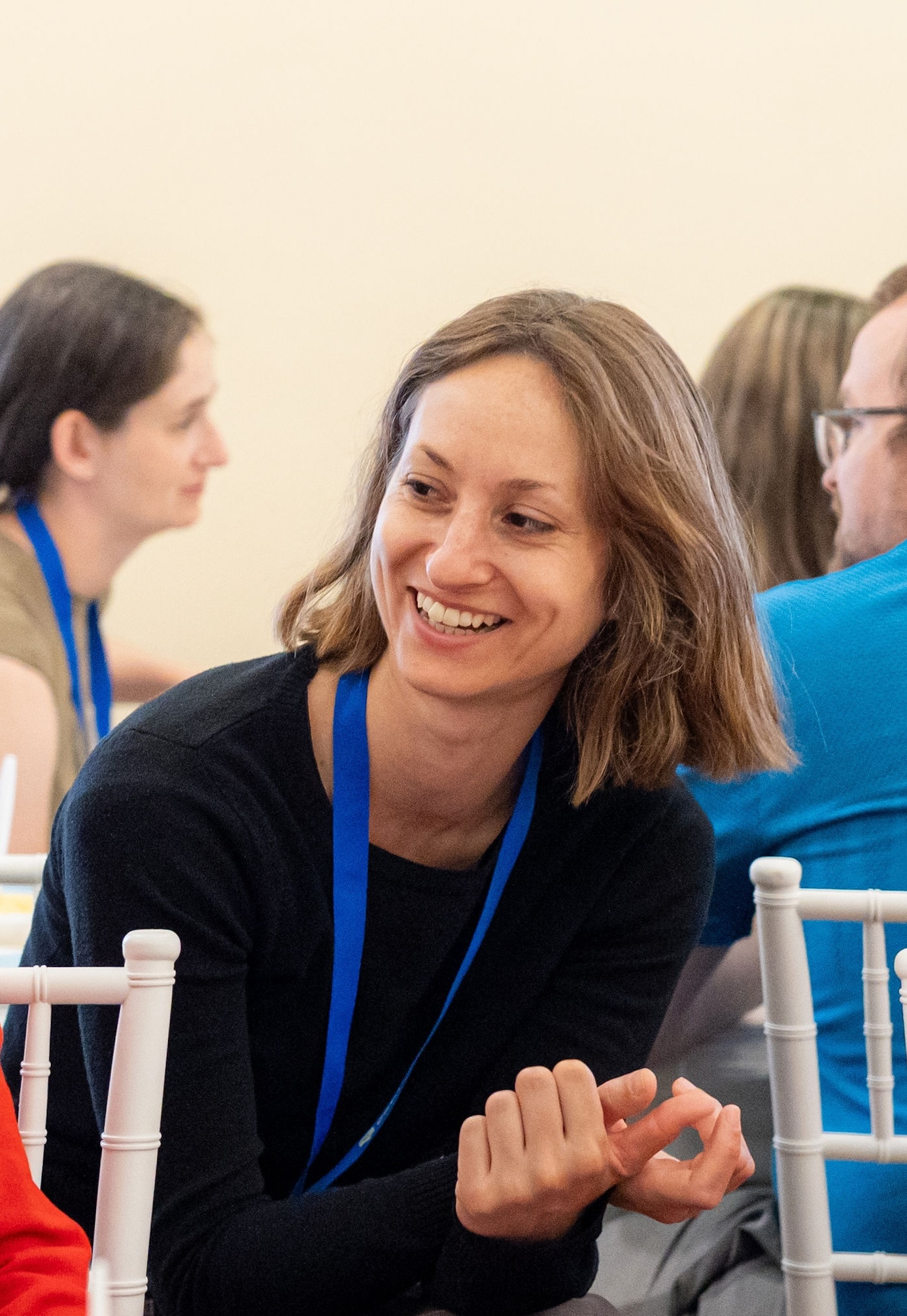 A smiling woman with shoulder-length light brown hair sits at a table during an event, wearing a black long-sleeved top and a blue lanyard. She is leaning forward in mid-conversation with her hands casually resting near her lap, surrounded by other attendees in a brightly lit indoor setting.