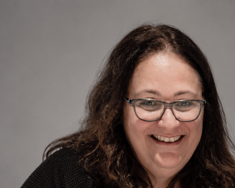 A smiling woman with glasses and dark, wavy shoulder-length hair, wearing a black sweater and an ID badge on a blue lanyard, seated at a white table against a gray background.