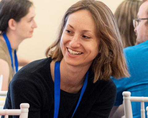 A smiling woman with shoulder-length light brown hair sits at a table during an event, wearing a black long-sleeved top and a blue lanyard. She is leaning forward in mid-conversation with her hands casually resting near her lap, surrounded by other attendees in a brightly lit indoor setting.
