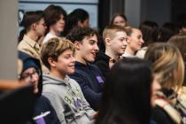 A group of young students sitting in an audience, smiling and listening attentively during a presentation or seminar.
